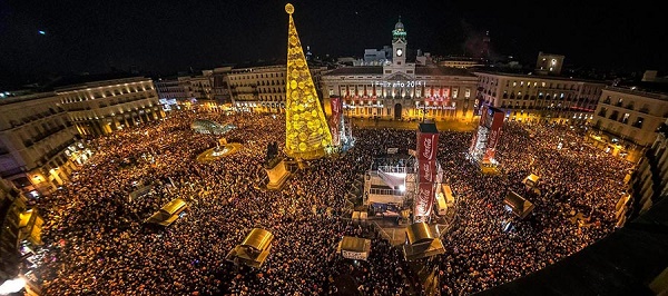 Tömeg a Puerta del Sol téren, Madridban, éjszaka, szilveszter napján. Nagy, sárga karácsonyfa, háttérben épület. Ünnepi hangulat, kivilágított tér. 