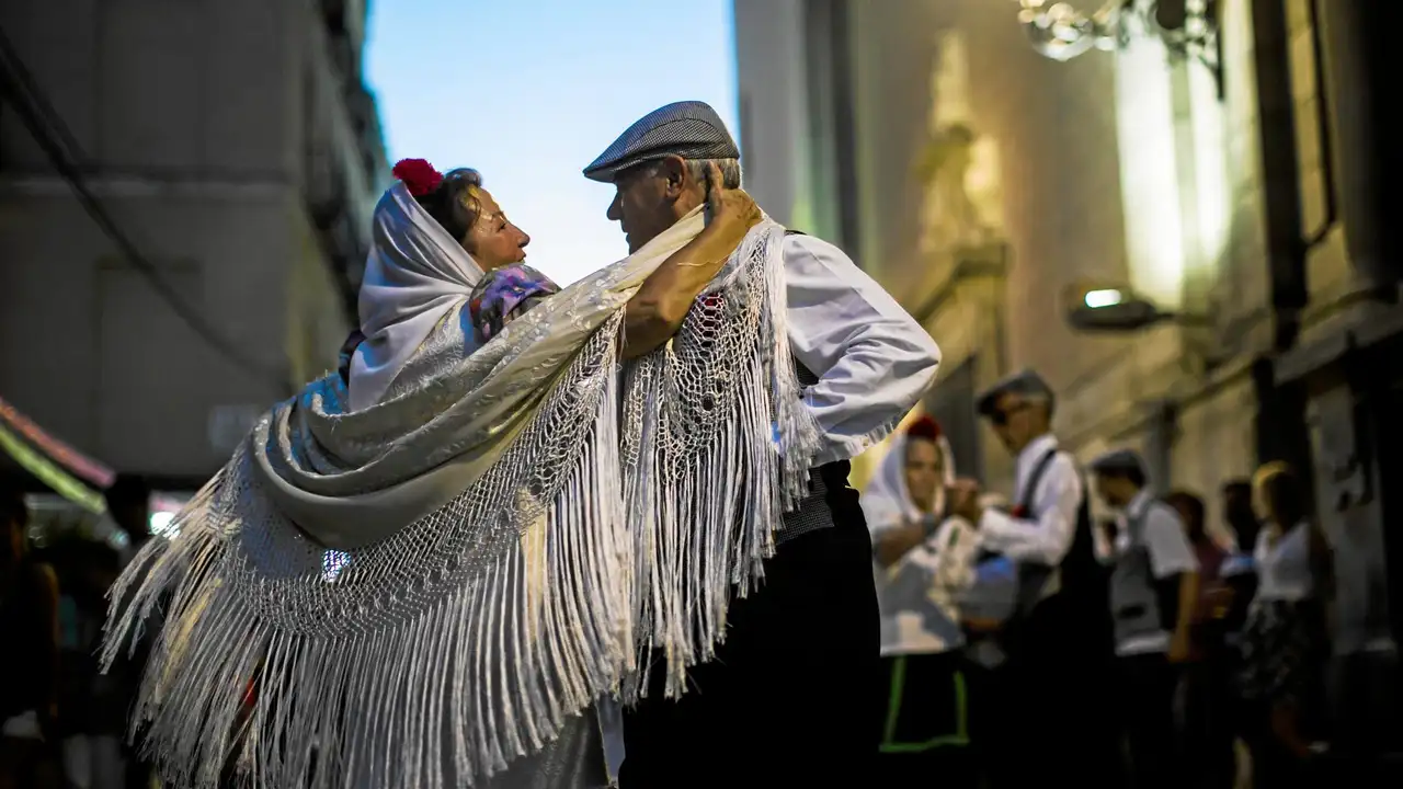 Idős pár tradicionális öltözetben táncol egy madridi parkban, háttérben egy nagy, klasszikus épület. Napfényes, derűs hangulat.