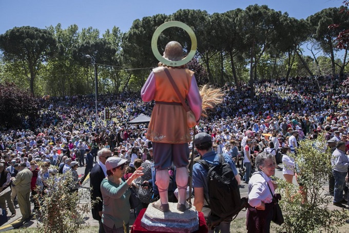 Madrid védőszentjének szobra, Földműves Szent Izidor áll szemben a tömeggel egy napsütötte parkban, háttérben zöldellő fák. Sokan kalapot viselnek, és vidám a hangulat.