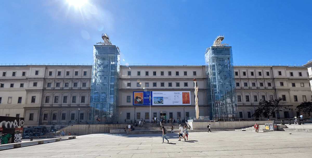 Reina Sofia múzeum Madridban, Spanyolországban. Széles, neoklasszikus épület üvegliftekkel, szobor elöl. Tiszta, kék ég, emberek sétálnak a tér kövén. Reklámtáblák a homlokzaton.