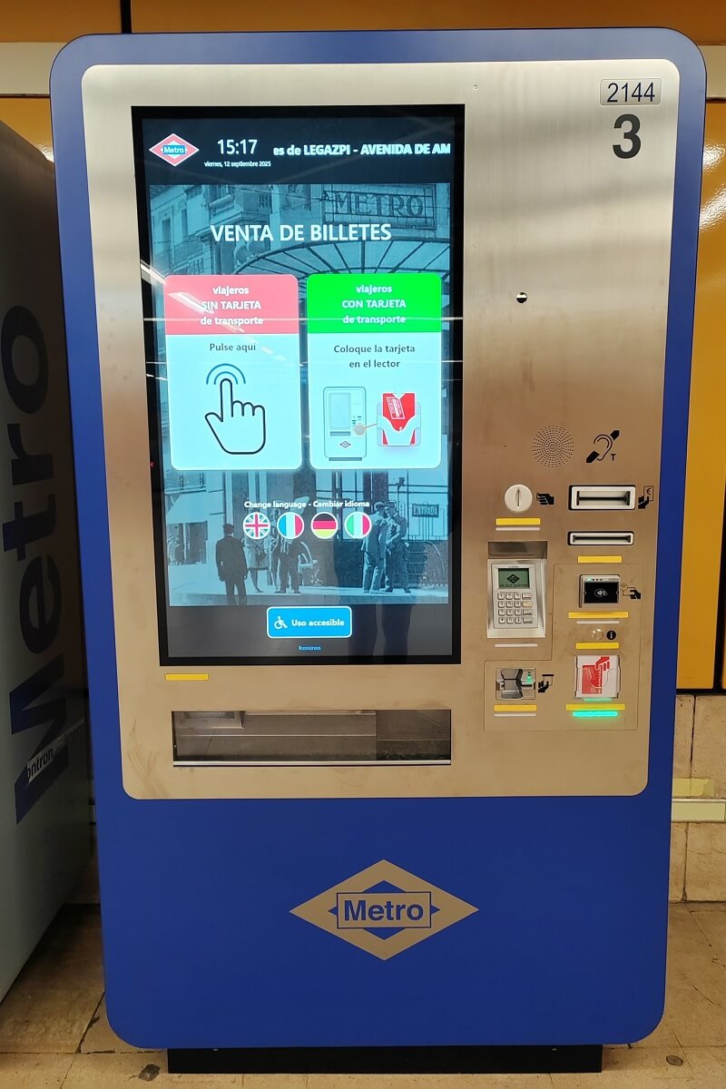 Kék metrójegy automata Madrid metró logóval. Érintőképernyőn jegyvásárlási opciók, nyelvek. Digitális kijelző és gombok láthatók.