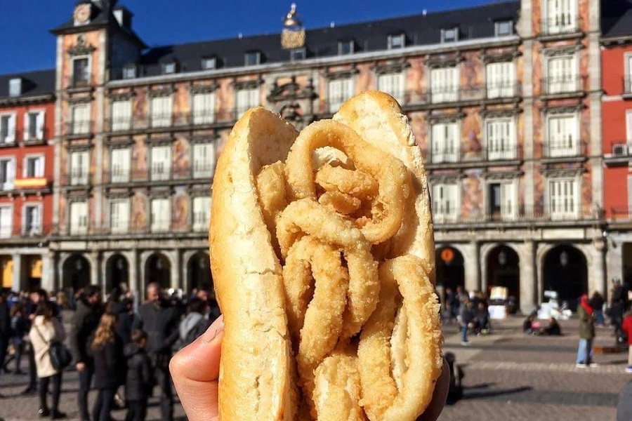 Madrid főtere, színes homlokzatú épülettel, előtérben valaki egy tintahalkarikás szendvicset mutat