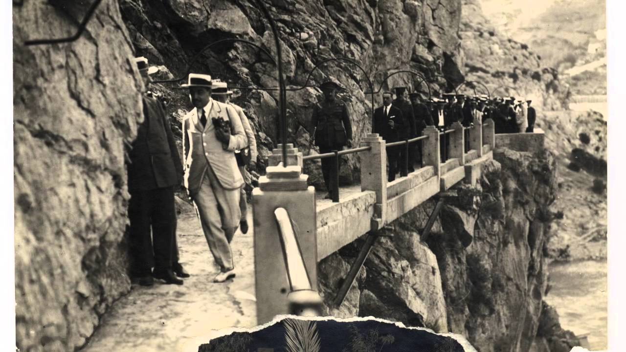 Emberek sétálnak egy szűk hegyi ösvényen, sziklafal mellett. Férfiak formális öltözetben, a hangulat feszültséget kelt. Retro stílus.