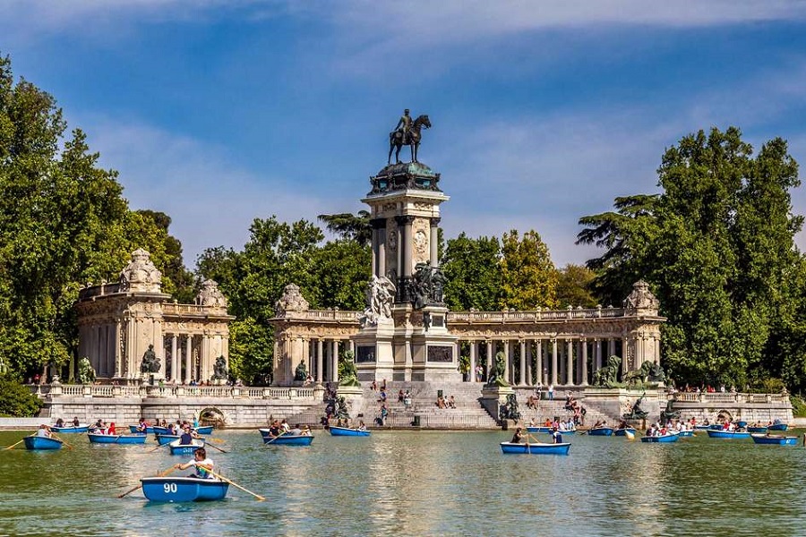 Madridi Retiro Park csonakázótóval, ahol éppen sokan csónakáznak, háttérben züld fák, kék ég és egy emlékmű, egy király lovasszobrával.