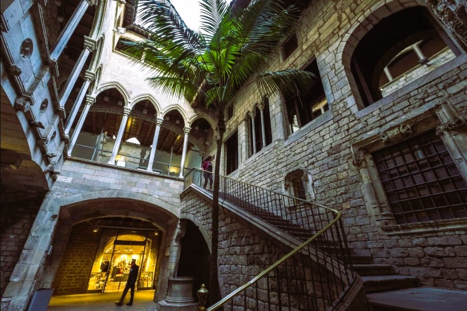 Két ember a barcelónai Picaso múzeum történelmi épületének kőudvarában, íves ablakokkal. Egyikük felmegy a lépcsőn. Bent meleg fények, középen pálmafa.