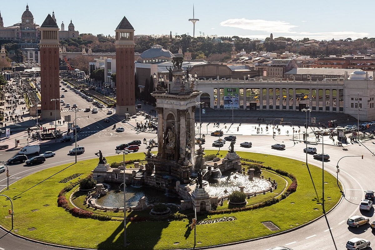 Forgalmas Placa d'Espanya tér körforgaloma, közepén szobor, háttérben két magas torony, városi táj. Felhős égbolt, nyüzsgő közlekedés és épületek.