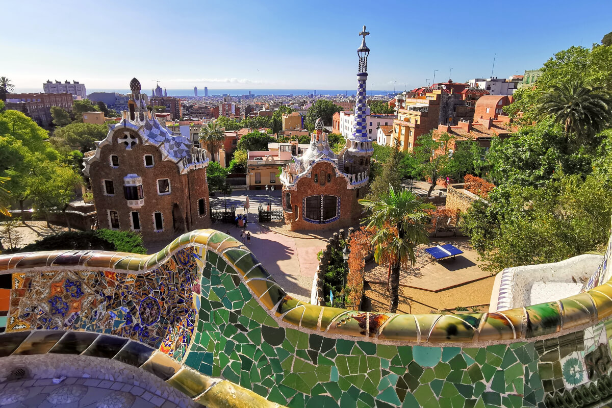 Gaudí Park Güell Barcelona. Színes mozaikos falak, egyedi épületek, zöld pálmafák, kék ég. Városi panoráma, nyugodt, derűs hangulat.