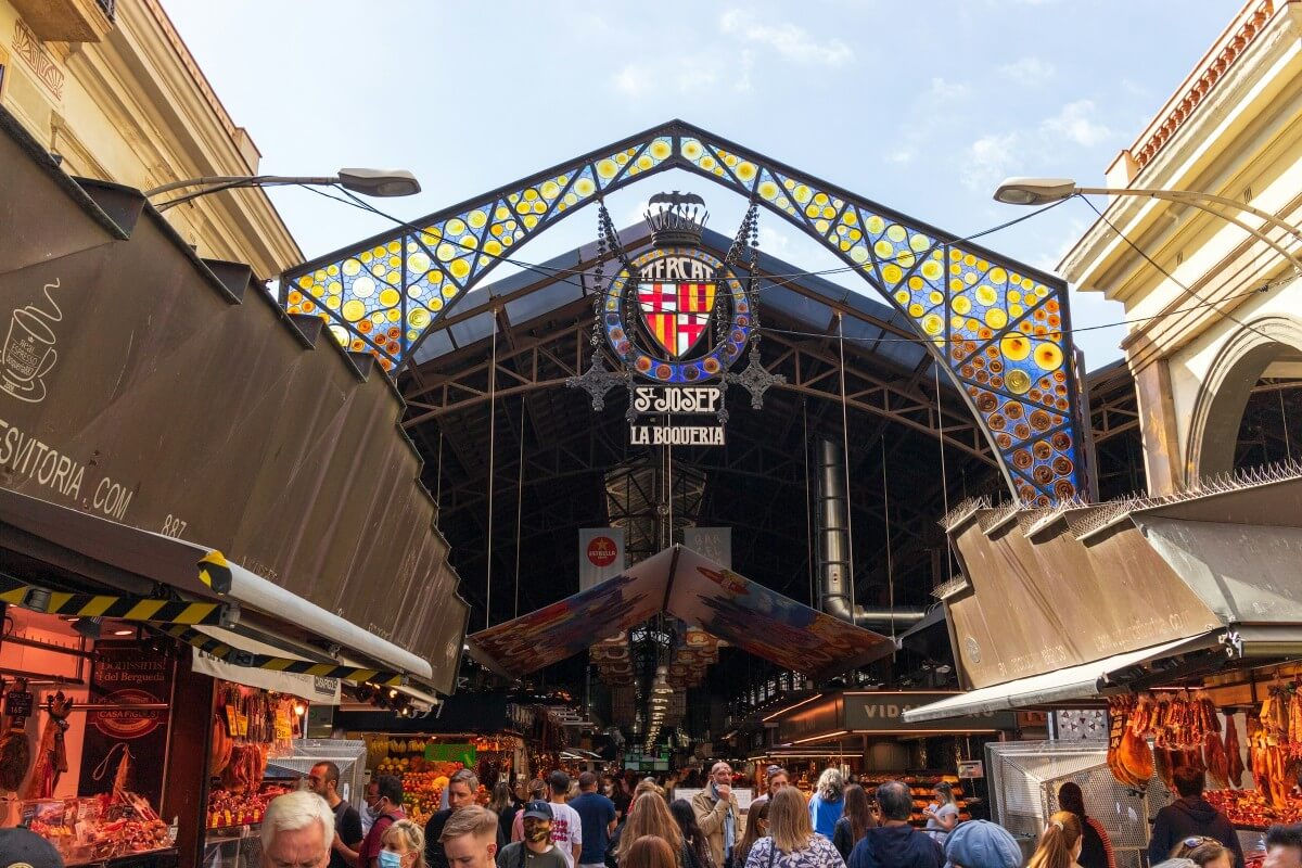 Színes mozaikokkal díszített piaci bejárat, La Boqueria felirattal. Emberek vásárolnak és sétálnak, élénk, nyüzsgő hangulat.