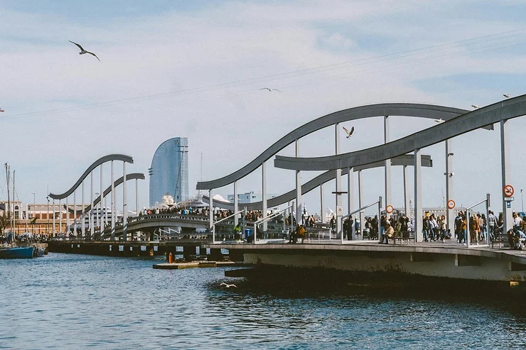 Rambla de Mar híd Barcelonában emberekkel, háttérben a W Barcelona hotel. Kék ég, madarak repülnek, nyugodt, tengerparti hangulat.
