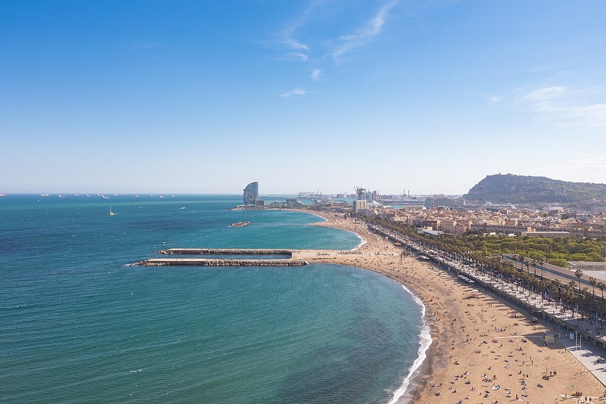 Tengerparti panoráma Barcelonában, kék ég, homokos Barceloneta strand, emberek napoznak, távolban felhőkarcoló és hajók. Szellős, nyári hangulat.