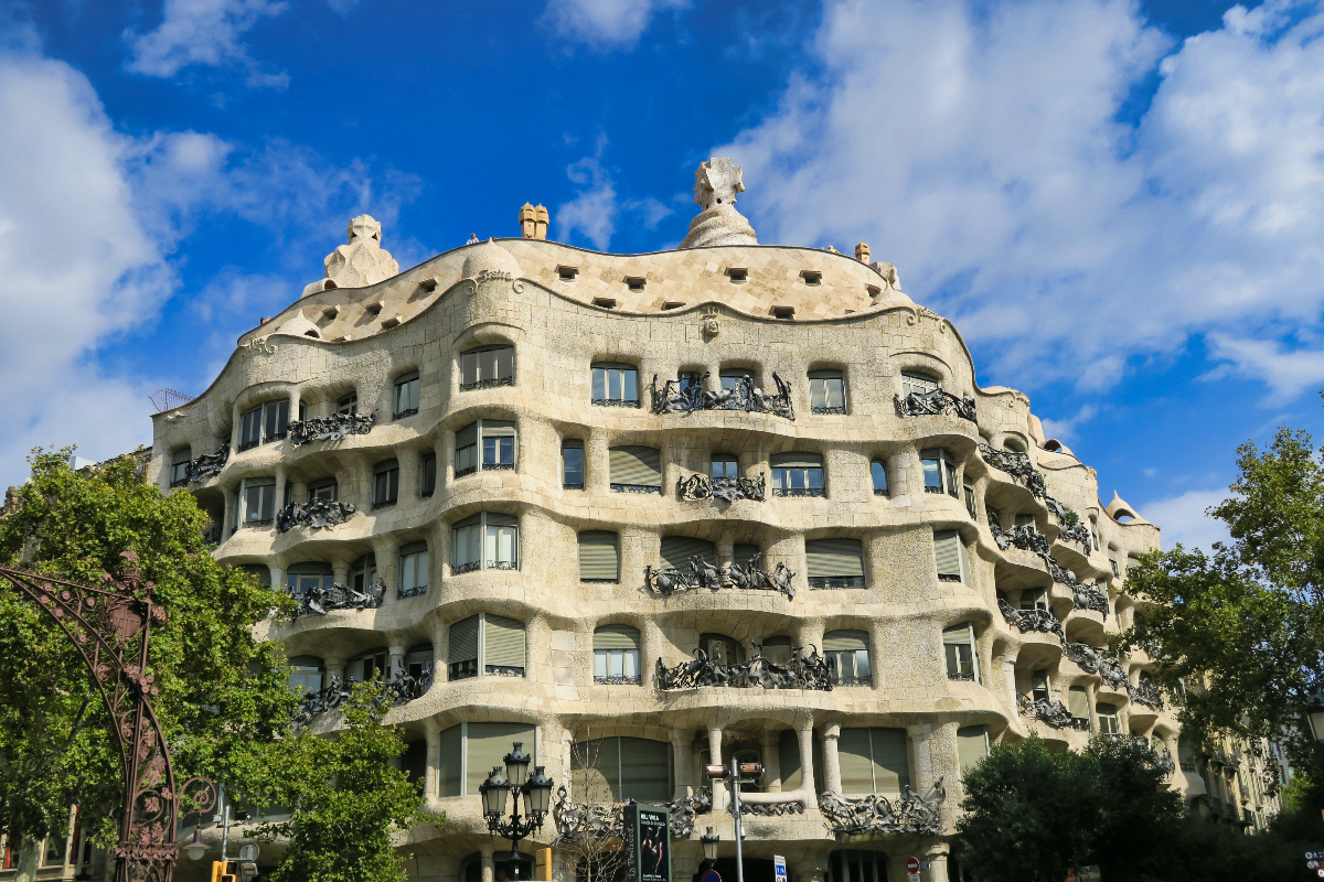 Hullámos homlokzatú épület világos kőből, kovácsoltvas erkélyekkel. Gaudí Pedrera háza Barcelonában. Kék ég háttérben, napfényes, modernista stílus.