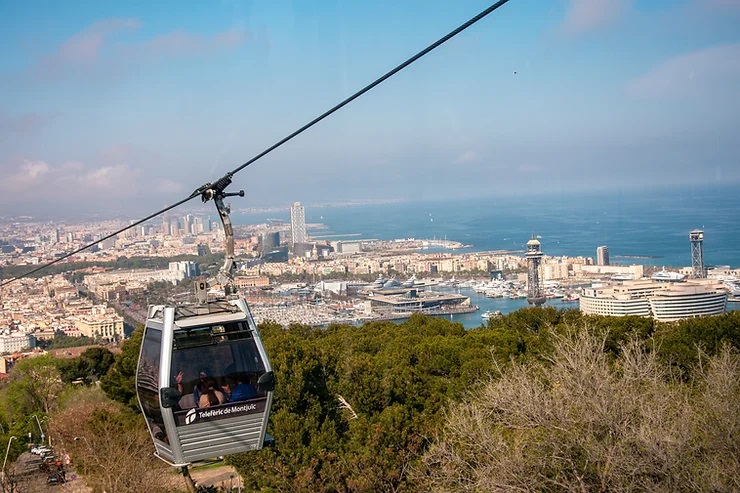 Kötélpályás kabin, Teleféric libegő Barcelonában, háttérben a város és a tenger. Napos idő, a kabin oldalán "Telefèric de Montjuïc" felirat látható.