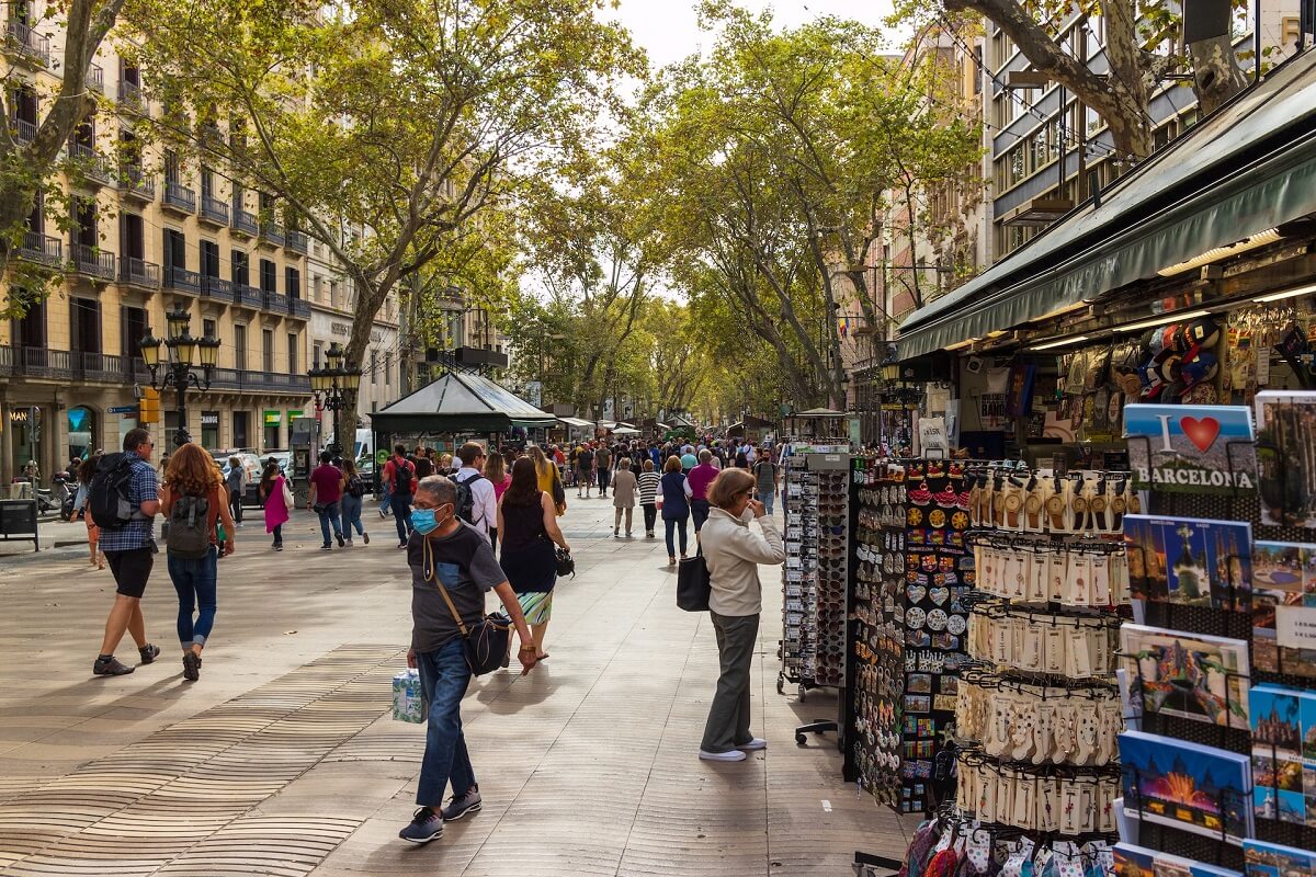 Forgalmas utca Barcelona szuvenírboltjával. Emberek sétálnak, árnyas fák alatt. Képeslapok, „I ❤ Barcelona” felirattal.