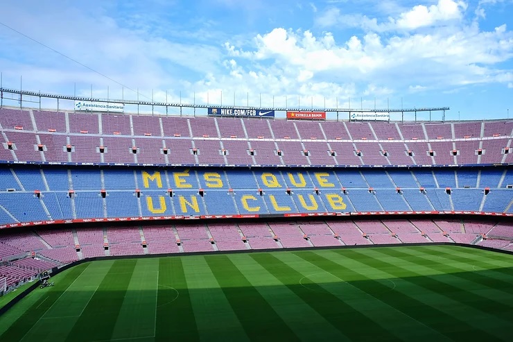 Üres Camp Nou stadion belső tere kék és piros széksorokkal. Középütt sárga felirat: MES QUE UN CLUB. Zöld gyep és kék ég látható.