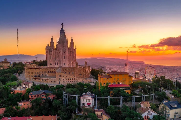 Szent Szív templom a domb tetején naplementében Barcelonában, körülötte házak, fák. Az ég narancssárga és lila, nyugodt hangulat.
