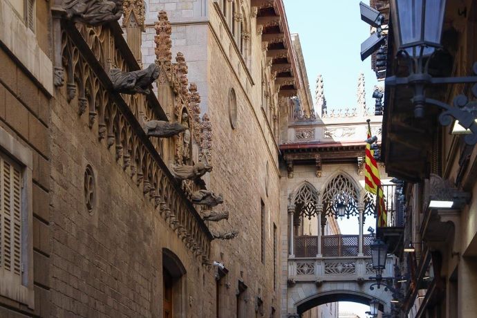 Szűk utcán faragott kő épületek, középkori stílusú csúcsíves híd köti össze őket Barcelona Gótikus negyedében. Jobbról piros-sárga zászló lóg, nyugodt légkör.