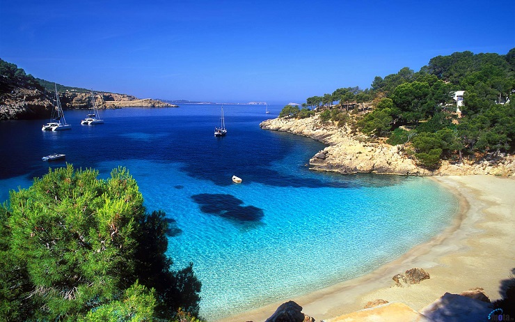 Ibiza, tengeröböl homokos parttal, kék vízzel. Vitorlások a távolban, buja zöld növényzet a háttérben. Nyugodt és békés hangulat.