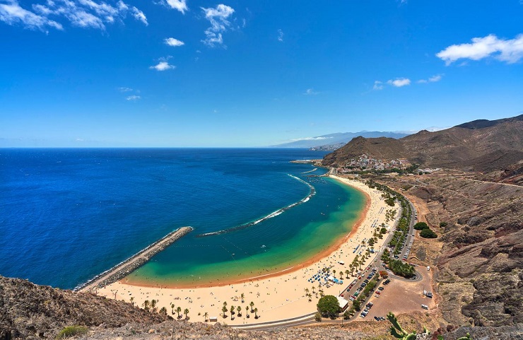 Tenerife, homokos tengerpart kék vízzel, pálmafákkal és hegyekkel a háttérben. Napos idő, tiszta égbolt, nyugodt, pihentető hangulat.