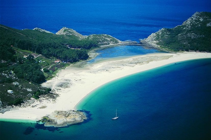Tengerparti táj kilátással kék vízre, fehér homokos stranddal, zöld hegyekkel a háttérben. Egy vitorlás a vízen, nyugalmas hangulat.