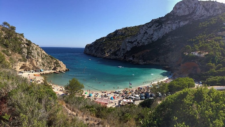 Tengerparti öböl Alicante mellett, strandolók napoznak és úsznak a türkizkék vízben. Zöld dombok és sziklák a háttérben. Napos, nyárias hangulat.
