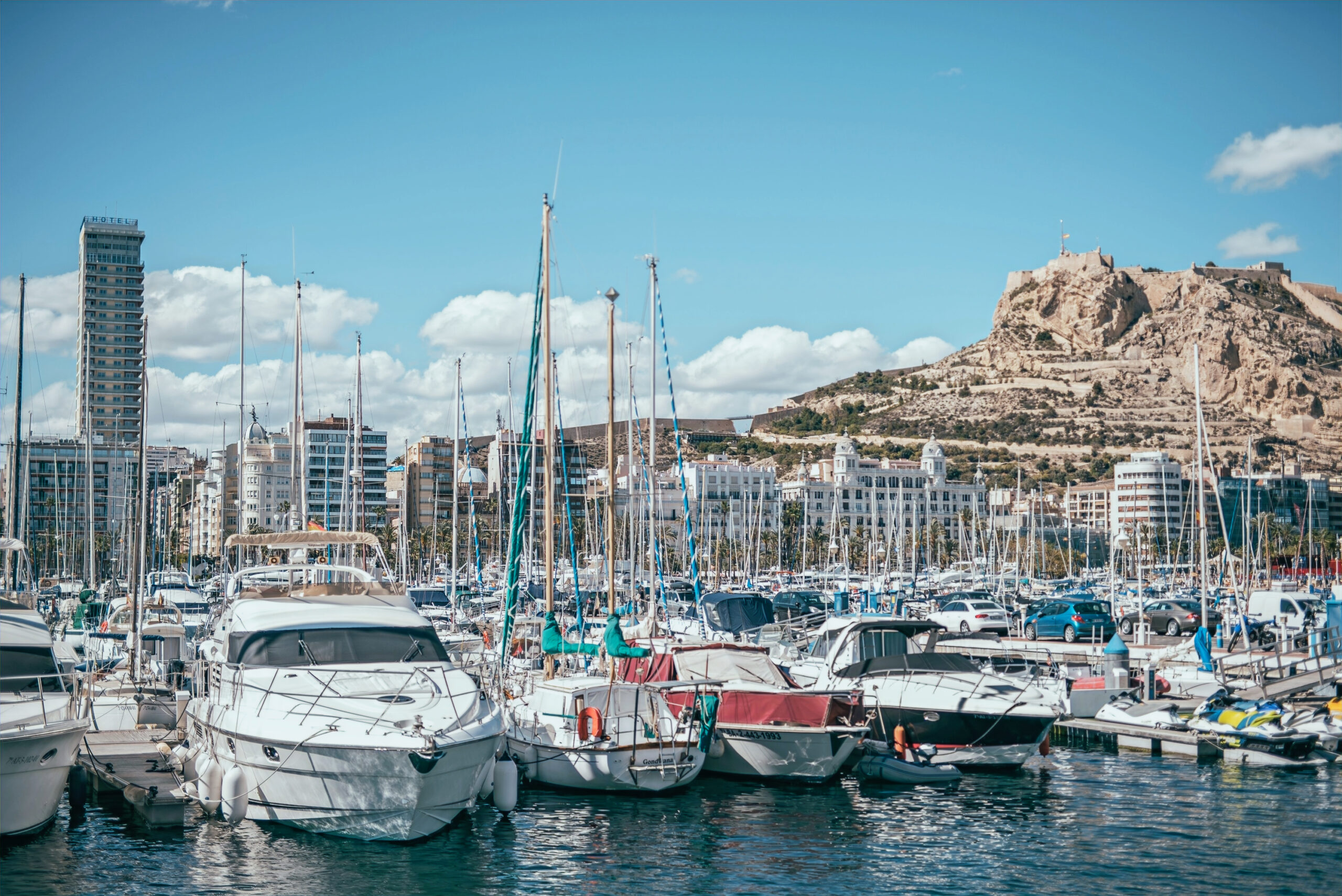 Alicante látnivaló, kikötő nagyon sok hajóval és vitorlással, háttérben a Santa Barbara vár.