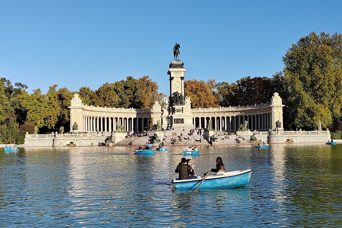 Emberek csónakáznak a Retiro Park taván, a háttérben a monumentális XII. Alfonz emlékmű oszlopsoraival és lovas szobrával – Madrid látnivalók.