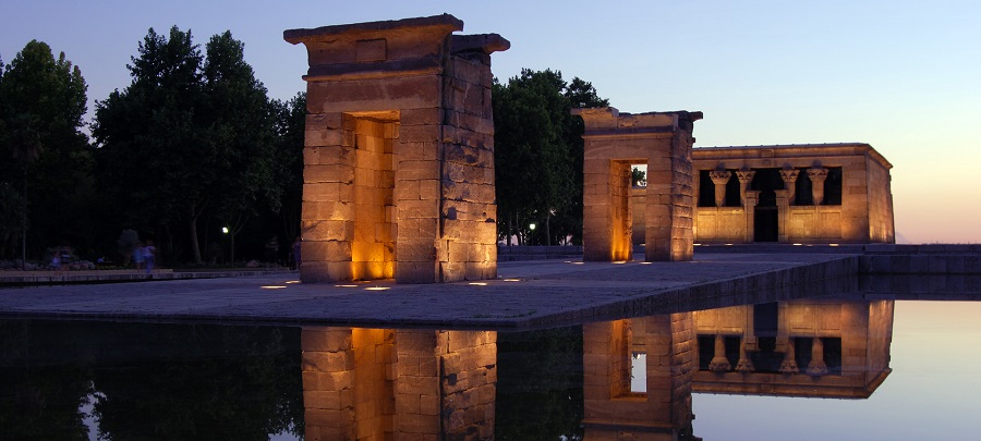 Templo de Debod ősi egyiptomi templom este, sárgán megvilágított kőépítmény, víztükröződéssel. Körülötte fák, égbolt halványlila és kék. Madrid egyik látnivalója.