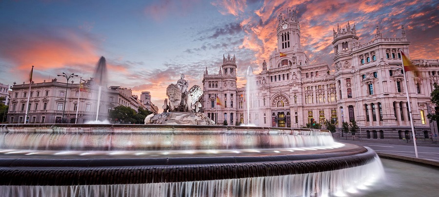 Cibeles-kút Madridban naplementében. Előtérben szökőkút, háttérben nagy épület. Égbolt narancs-rózsaszín, hangulat nyugodt.