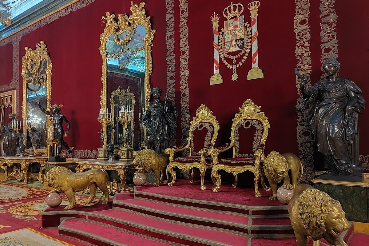A madridi Királyi Palota trónterme. Piros fal szobrokkal, aranyozott tükrök, trónszékek, oroszlánokkal. Elegáns és királyi atmoszféra, címer a falon.