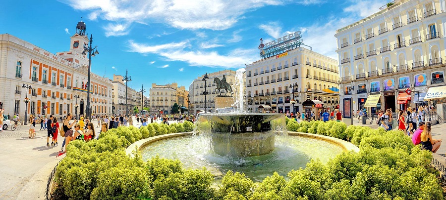 Puerta del Sol, ami forgalmas tér Madridban, emberekkel a szökőkút körül. Napos idő, kék ég. Környező történelmi épületek és üzletek színes táblákkal.
