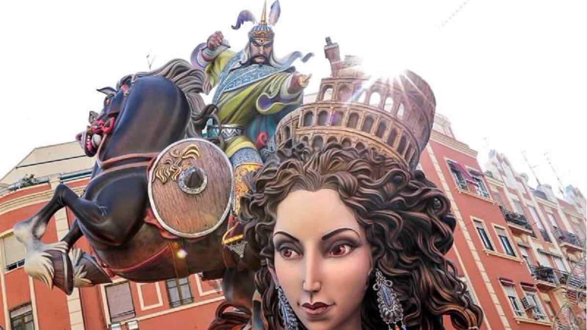 Las Fallas fesztivál Valencia, Spanyolország. Impozáns szobor: női fej és harcos lóháton, színes ruhában, pajzzsal. Hátul vörös épület, napfénytől ragyogó részletek. Ünnepi hangulat