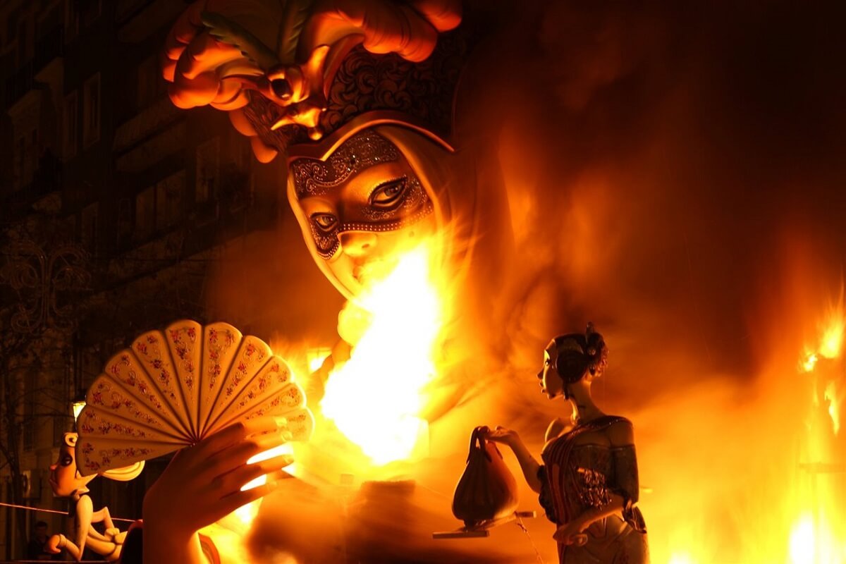 Monumentális maszkos figura lángokat fúj a valenciai Las Fallas fesztiválon, körülötte narancssárga fények és füst, drámai és misztikus hangulat.