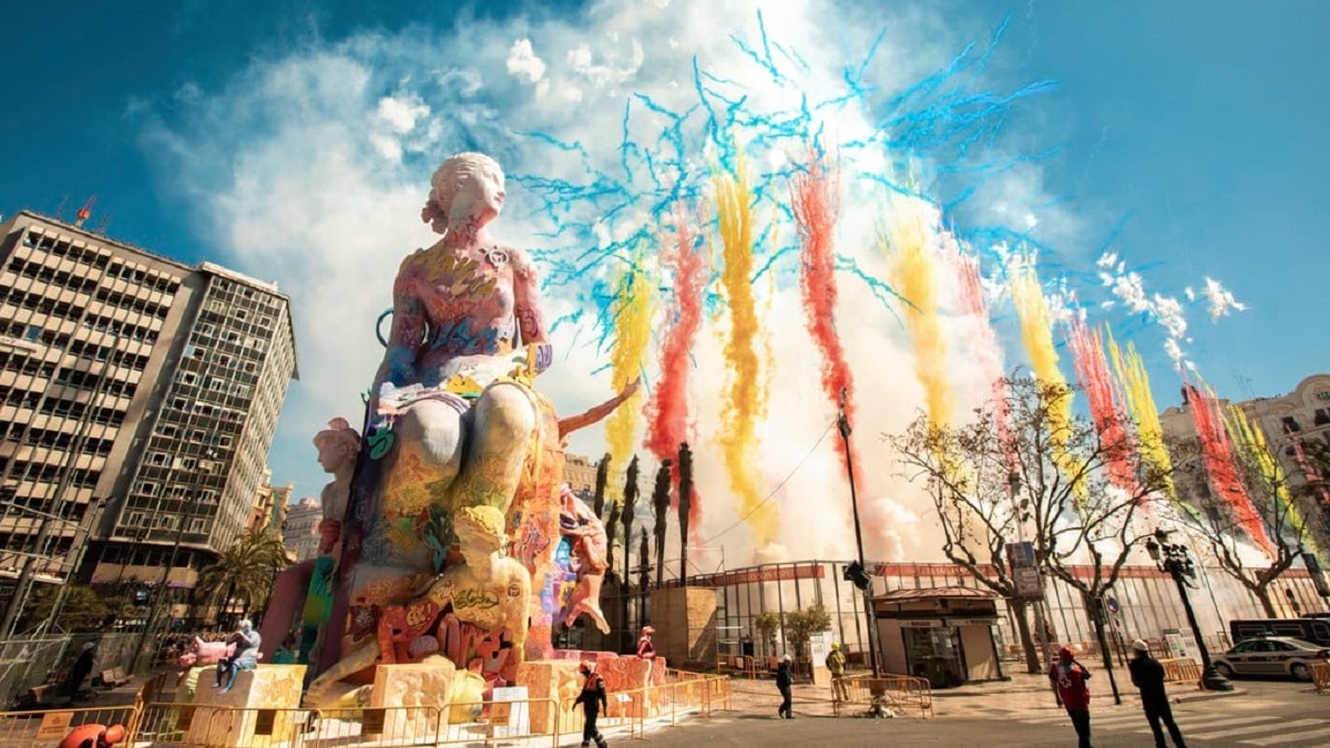 Színes szobor egy téren, háttérben színes füst és ünnepi hangulat. Kék ég, épületek, élénk színek. Emberek figyelnek a földről a Las Fallas valenciai fiestán.