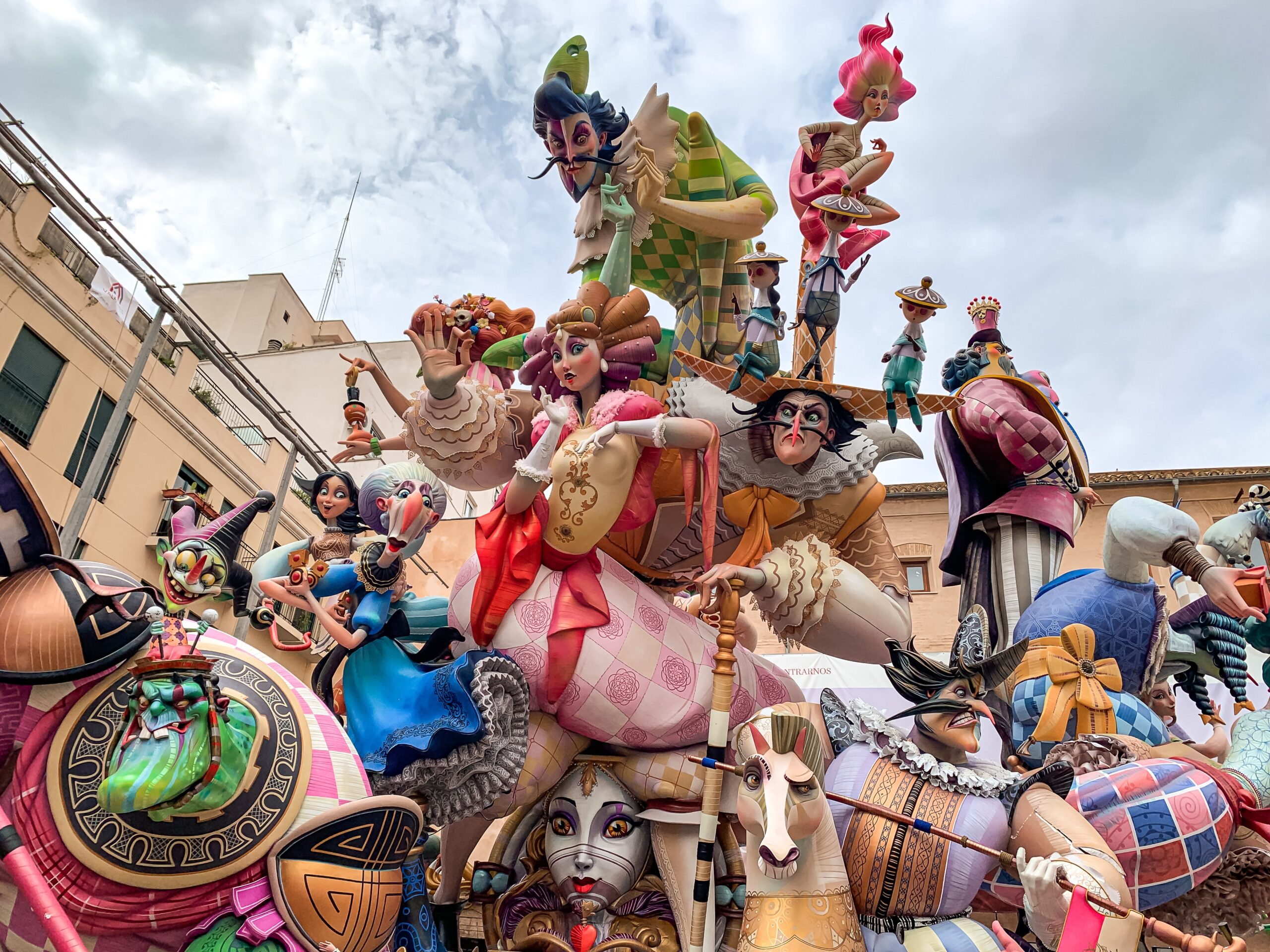 Las Fallas fesztivál Valenciában. Sokszínű, színes szobrok mesebeli karaktereket ábrázolnak egy városi tér hátterében, felhős éggel. Élettel teli, vidám hangulat.