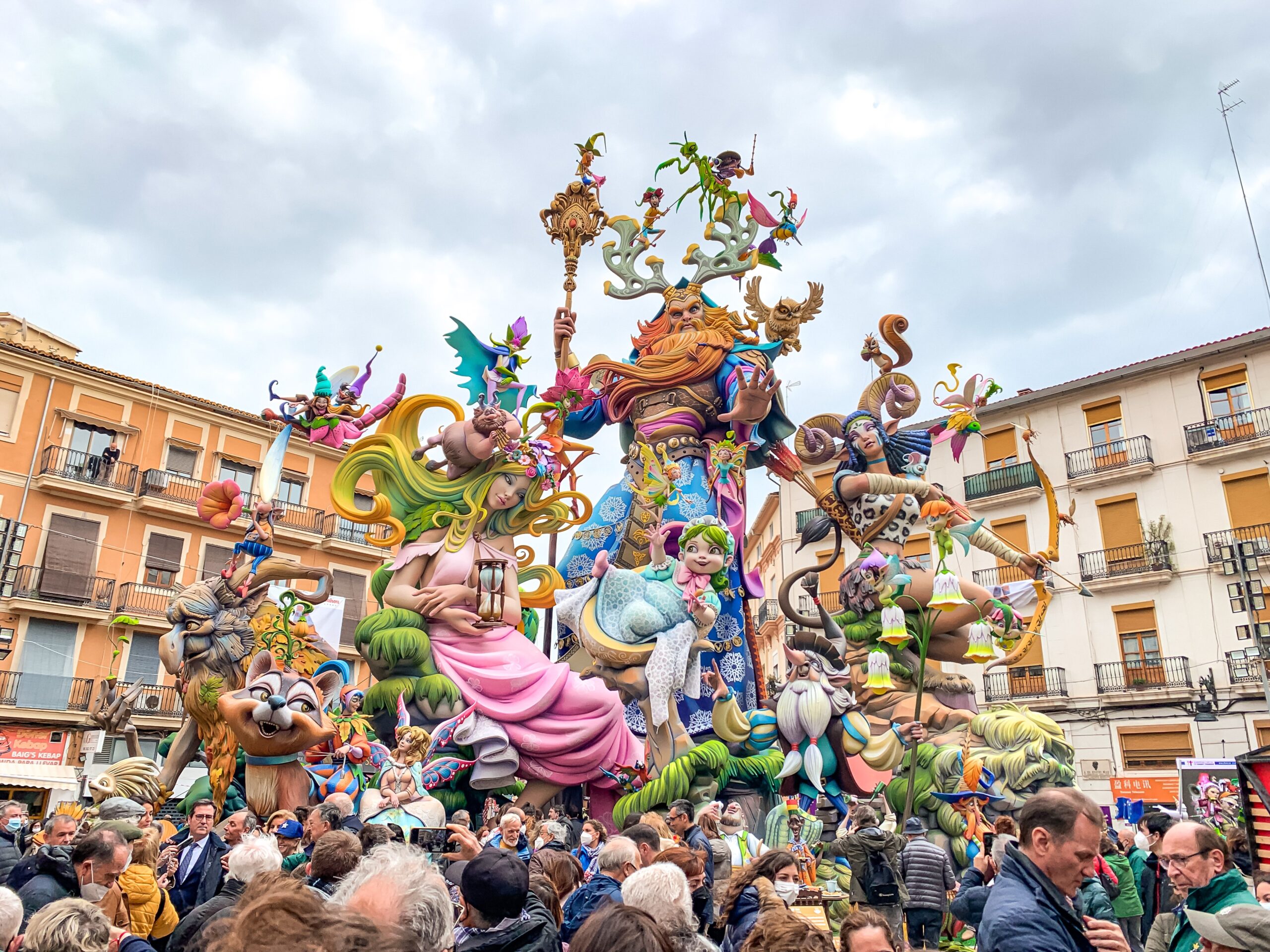 Las Fallas színes fesztivál szobor ábrázol mesealakokat, körülötte tömeg. Hátterében narancssárga épületek és felhős égbolt.