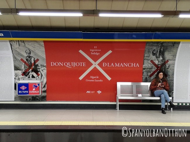 Madridi metróállomás poszterrel: Don Quijote vastagon piros plakáton. Jobbra ülő nő mobilozik. Fém padok, világos fények.