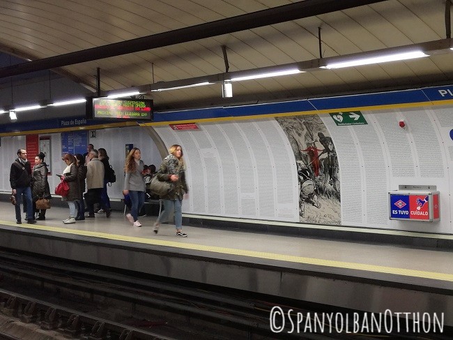 Emberek sétálnak egy madridi metróállomáson, a háttérben színes hirdetés, fehér fal, grafika és vészkijárat jel. Kényelmes hangulat.