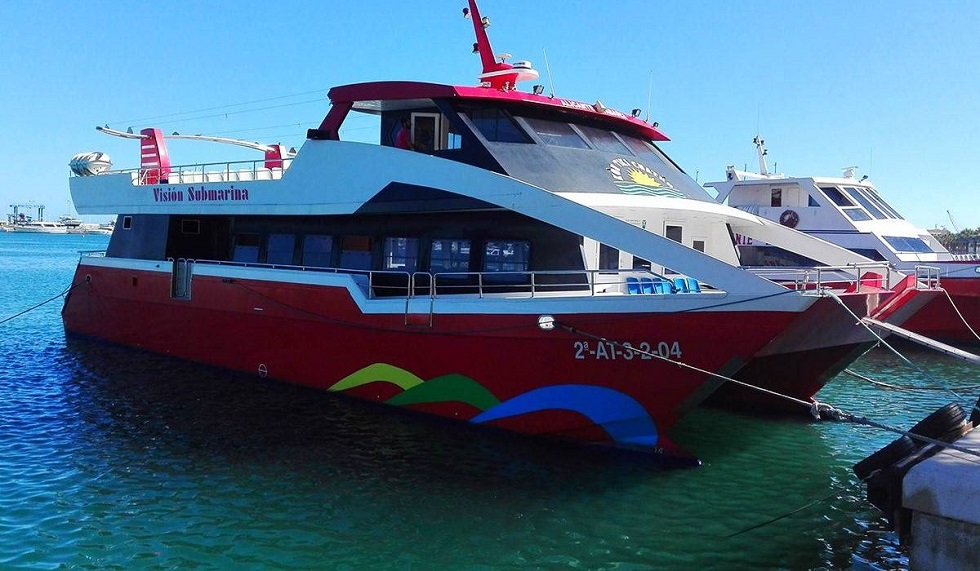 Alicante hajókirándulás katamaránnal Tabarca szigetre. Piros hajó a vízen "Visión Submarina" felirattal, kék ég alatt. Mellett egy másik hajó. Nyugodt kikötői hangulat.
