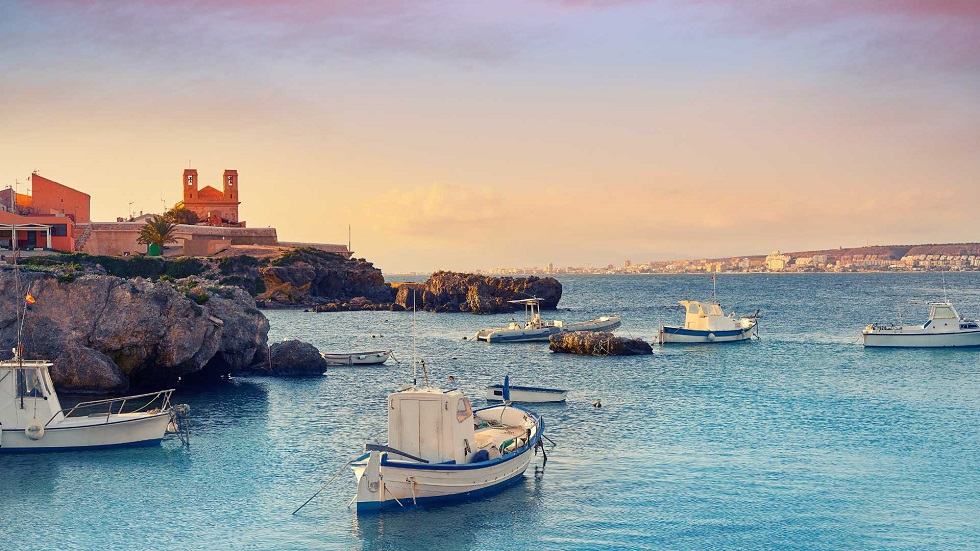 Alicante hajókirándulás Tabarca szigetére. Hajók horgonyoznak a kék tengeren egy sziklás part közelében, háttérben egy vörös épület. Naplemente meleg fényével vonja be a tájat.