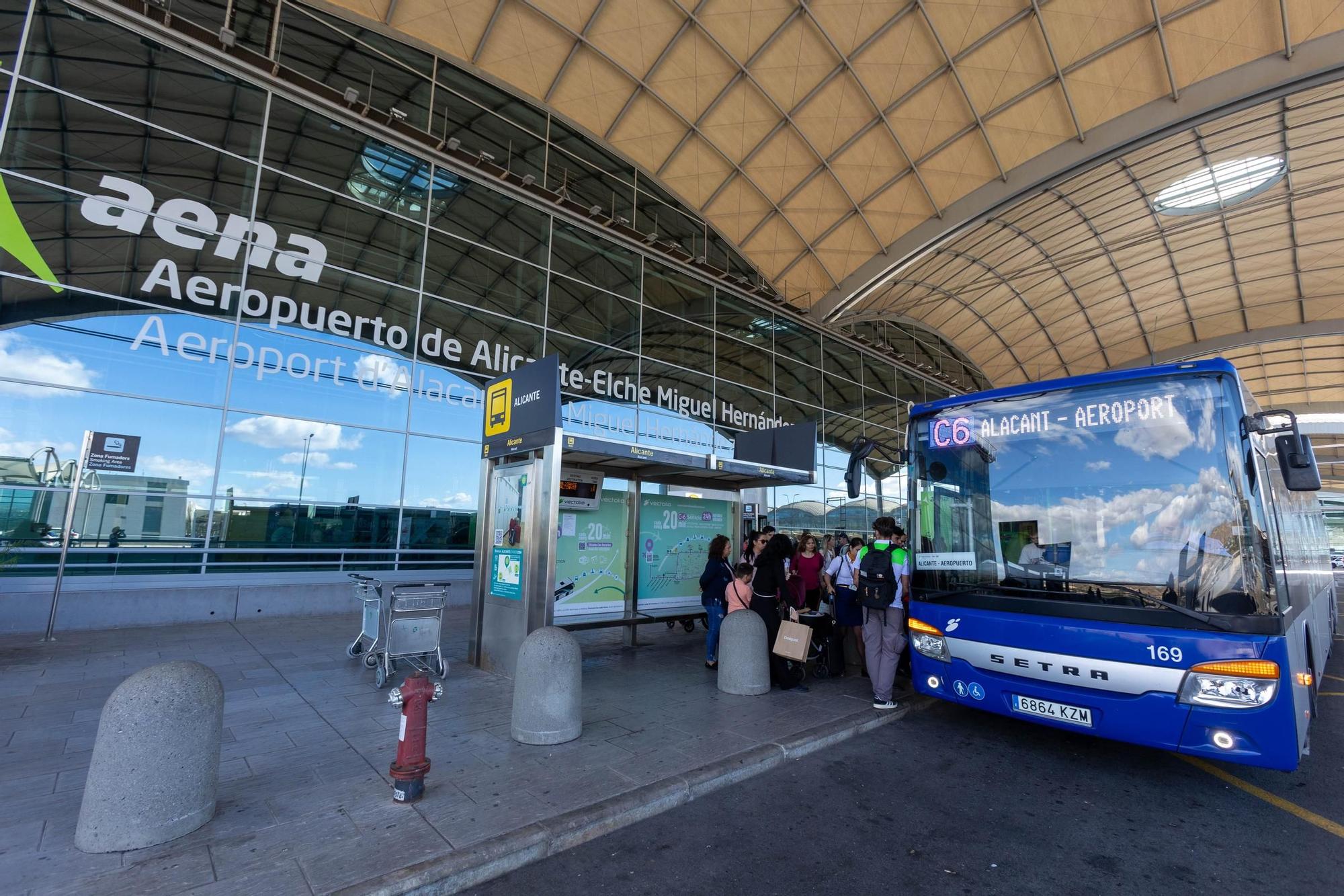 Alicante repülőtér bejárata ami előtt a C6 kék reptéri busz várakozik, és emberek akik felszállnak a buszra