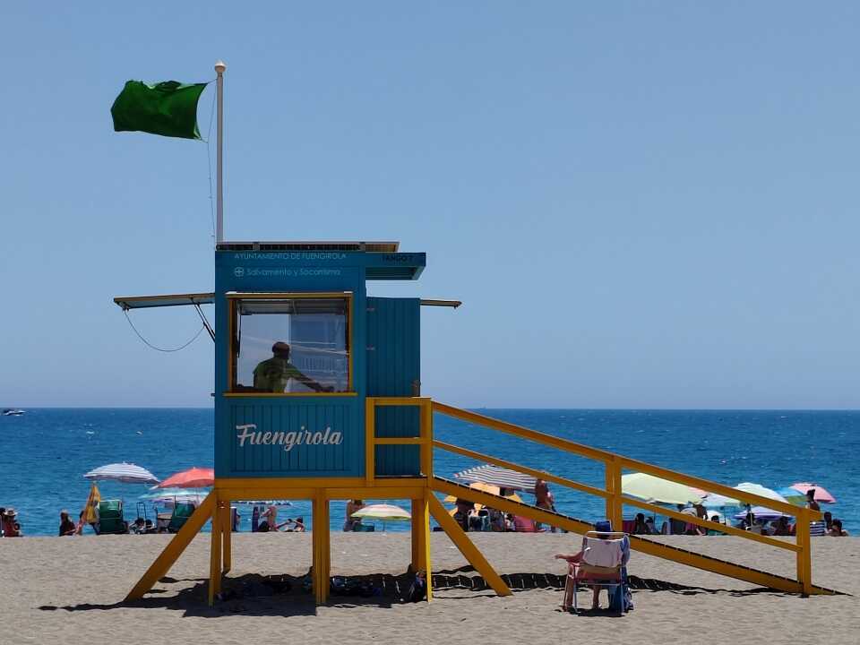 Fuengirola tengerpart a vizimentő bódéja, rajta zöld zászló, háttrében kék tenger és ég, és napozók