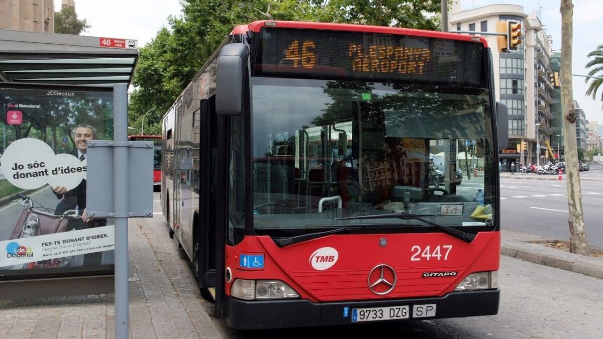 Piros busz Barcelonában, 46-os járat repülőtér felé. Hirdetőtábla közelében, zöld fák és modern épületek a háttérben.