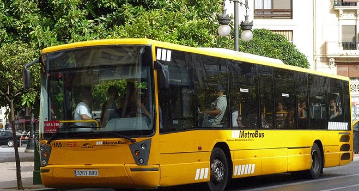 Sárga MetroBus az utcán, utasokkal Valenciában. Zöld fák a háttérben, napos idő. Felirat: "MetroBus".