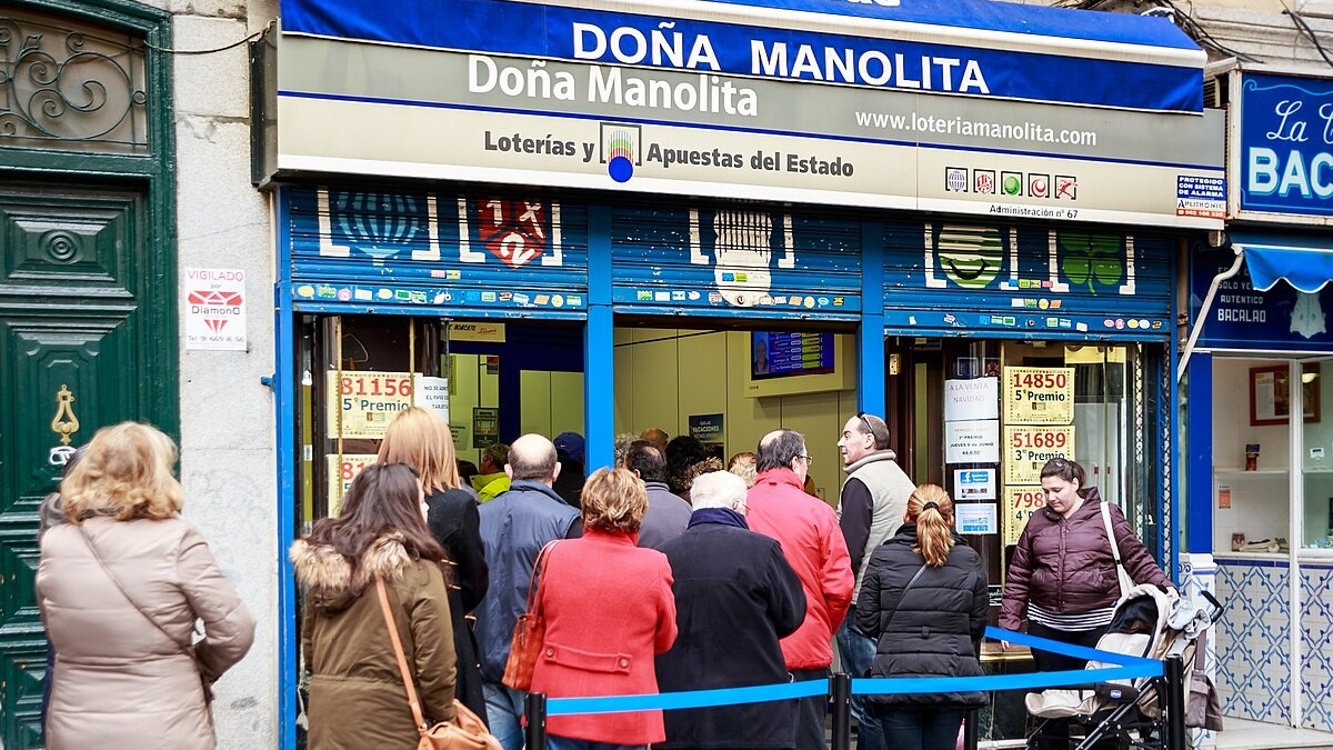 Emberek sorban állnak a Doña Manolita lottózó előtt Madridban. A háttérben épületek, a hangulat feszült, várakozó.