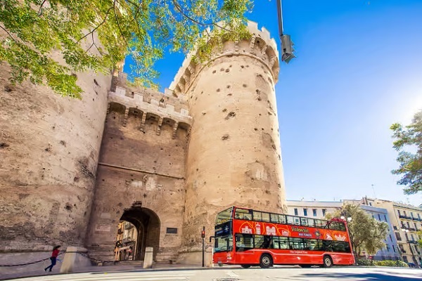 Piros emeletes busz halad Valenciában a Torres Serranos középkori vár kapuja előtt. Kék ég, napfény, kőtorony és fák a háttérben. Turistás hangulat.