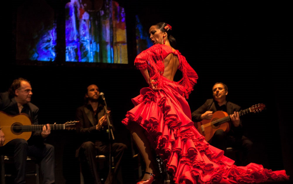 Flamenco táncos piros ruhában egy madridi tablaóban, háttérben gitárosok